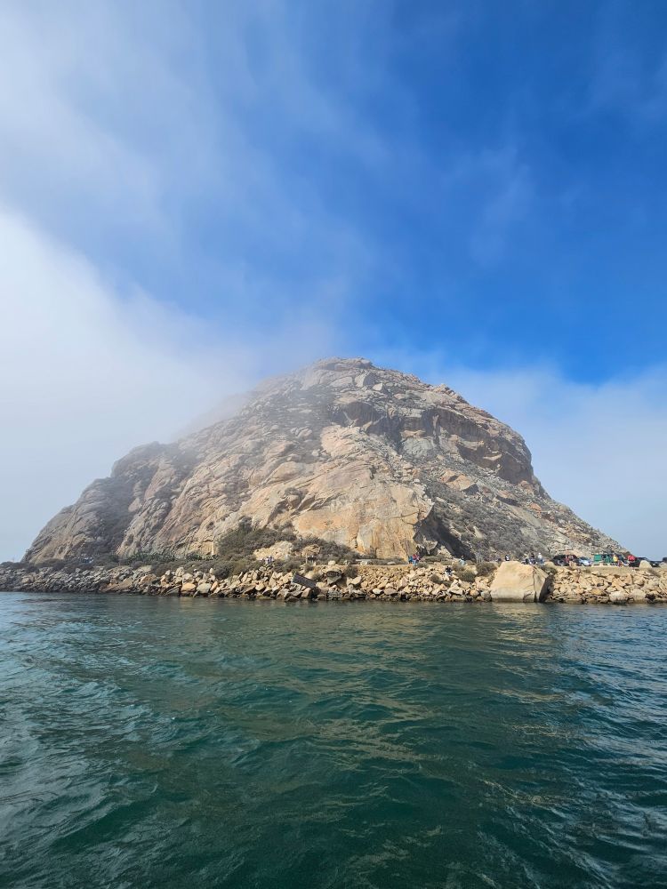 Morro Rock towers above the eponymous bay, which is a dark green color. Wisps of fog play around the shoulders of the mountain.