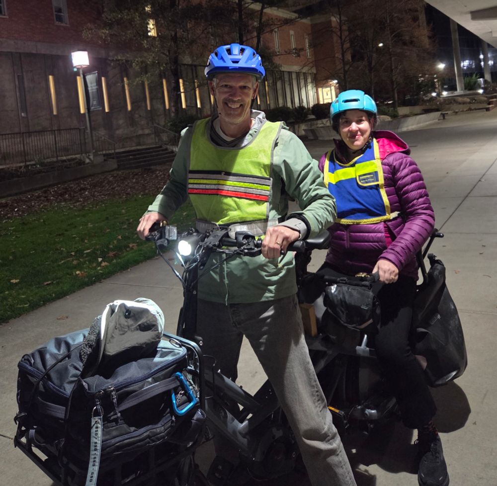 Dr Tom Albright stands over his cargo bike while Dr Tara Goddard sits on the rear seat of the bike. Their bags are piled in the front and panniers hang off the back rack. They are both bundled up and smiling.