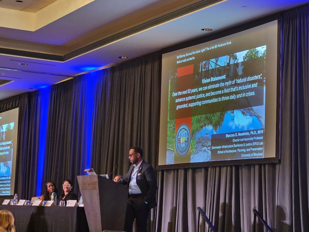 Dr Marccus Hendricks on stage in front of his vision statement about the need to center environmental justice in emergency management research and practice