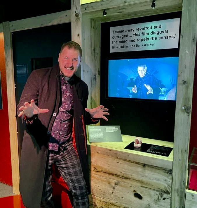 Smiling man pretending to be a vampire next to an exhibit about Dracula 