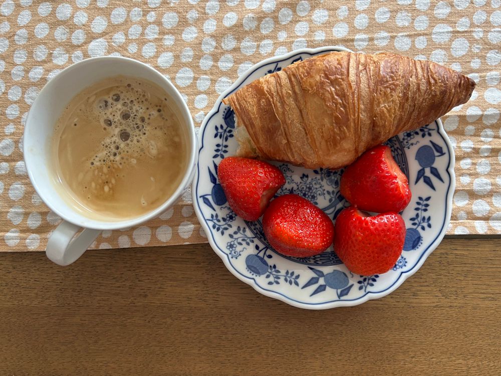 Auf einem Tisch steht eine Tasse mit Kaffee sowie einen Teller mit einem angebissenen Croissant und vier riesigen Erdbeeren