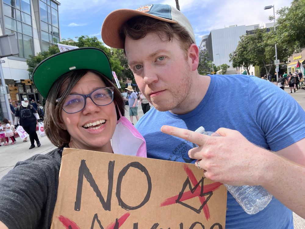 A woman with short brown hair and a baseball cap and glasses is standing next to a very cool man named Brennan. Brennan is wearing a baseball cap and blue shirt and it pointing to the woman. The woman is holding a cardboard sign that says “No Kings”. 