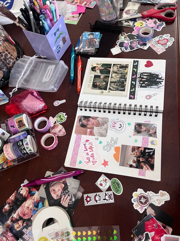 An open scrapbook lays on a table. There are stationary items (pens, washi tape, stickers) scattered. Across the table and in the scrapbook are pictures and stickers of members of the Kpop band Stray Kids. 