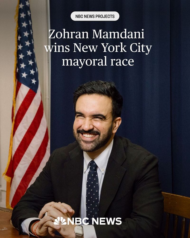 Zohran Mamdani Wins New York City Mayoral Race is over a smiling picture of Zohran Mamdani with his hands folded, elbows on the table. 
