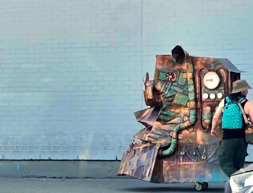 A contraption from Clay and Paper Theatre in Toronto is being rolled down the sidewalk beside a large white brick wall. There's a person in a hat and backpack turning behind them to look at something. 