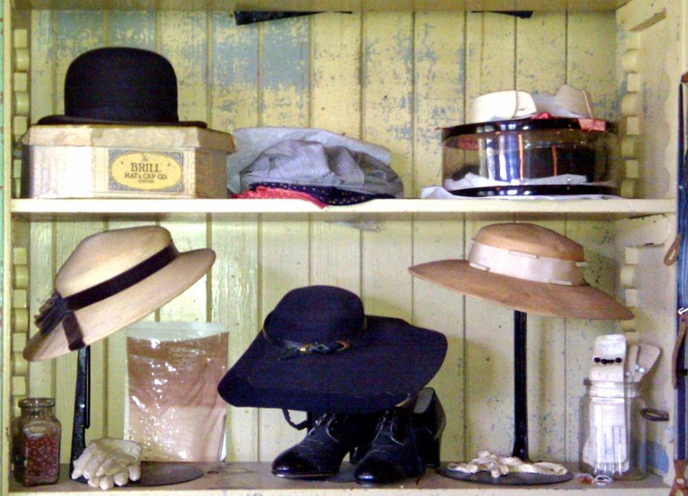 A two shelf display of assorted styles of vintage hats and caps. Men's above and women's below. The box under the bowler hat above reads BRILL Hat & Cap Co. There's also a flat cap and a round hat box with a mystery in it. Bottom shelf also has white gloves, jars of buttons and some old lace up black leather heeled booties, in addition to three brimmed hat with ribbons. 