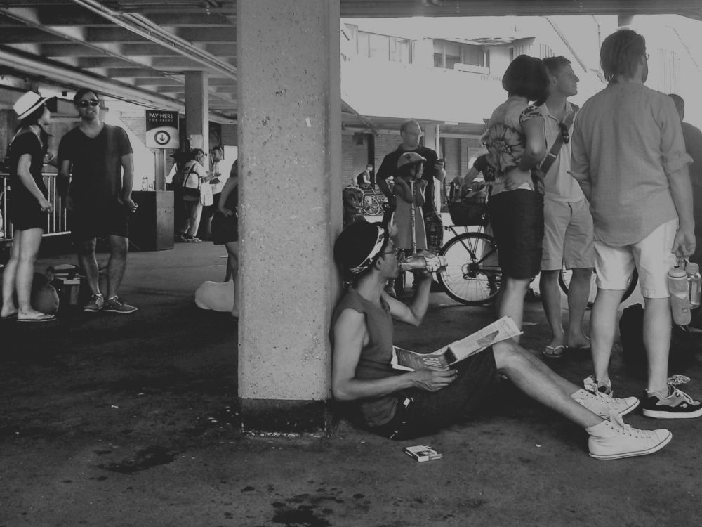People stand around at the ferry terminal in downtown Toronto waiting for a ferry to the Toronto Islands. Someone is sitting on the ground against a column drinking a soda and reading the paper. 