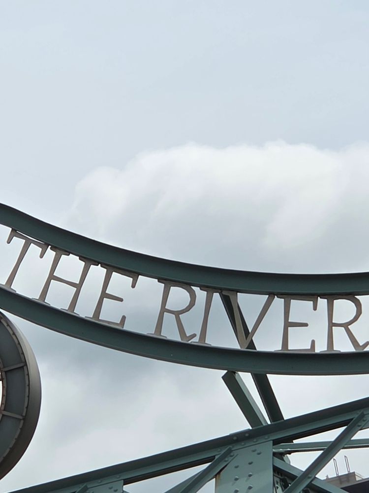 The words "THE RIVER" cropped from a quote spanning above the Riverside Bridge (Formerly Queen St. Viaduct) in Toronto. Some of the steel framework is visible under the bridge. A cloud looks like it's resting in the scoop made by the curve of the words. 