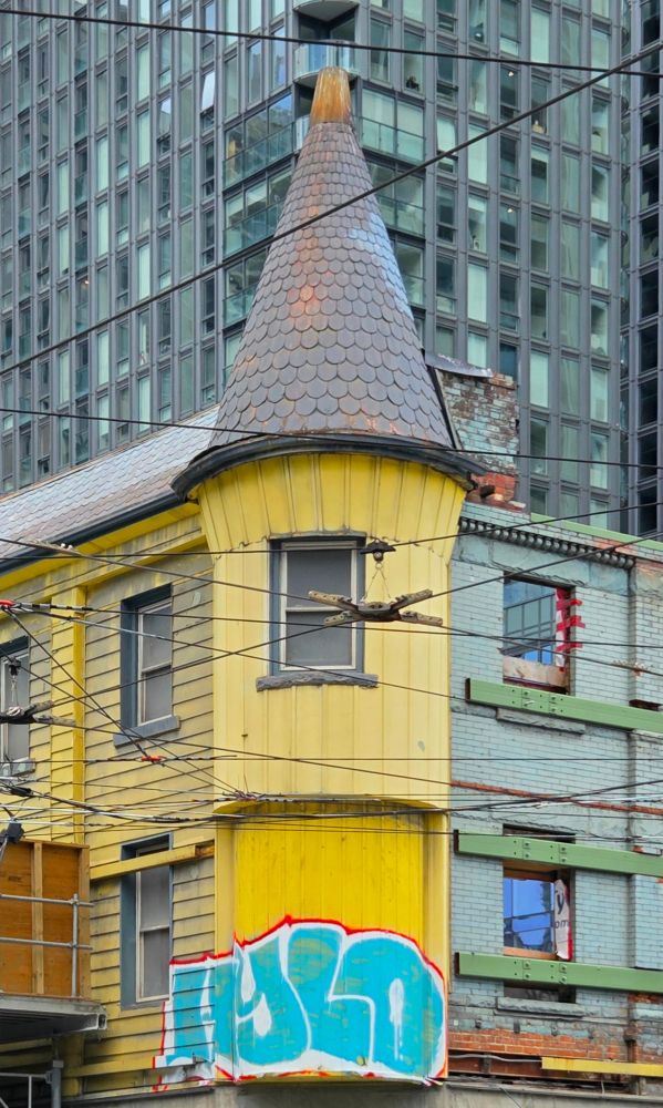 Two buildings at a corner in downtown Toronto. The one in the foreground is a bright yellow turreted heritage building, currently undergoing renovations. The siding has been removed on one side revealing the original brick wall. Someone has graffitied the word "HYLO" on the bottom of the turret. A wall of windows of a glass and steel condo looms high in the background. Streetcar wires criss-cross the foreground. 