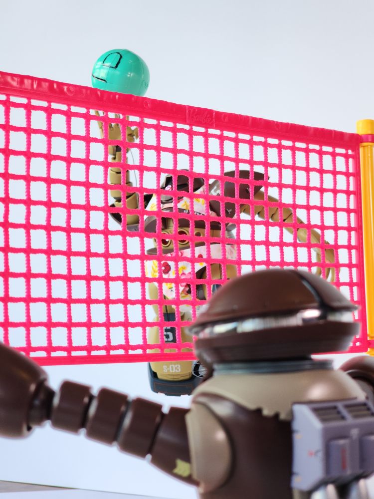 A photo from behind a HG Acguy who is attempting to block a shot from a HG Gogg who is mid-jump to spike a volleyball over a net. The HG Gogg is wearing a floral pink bikini.