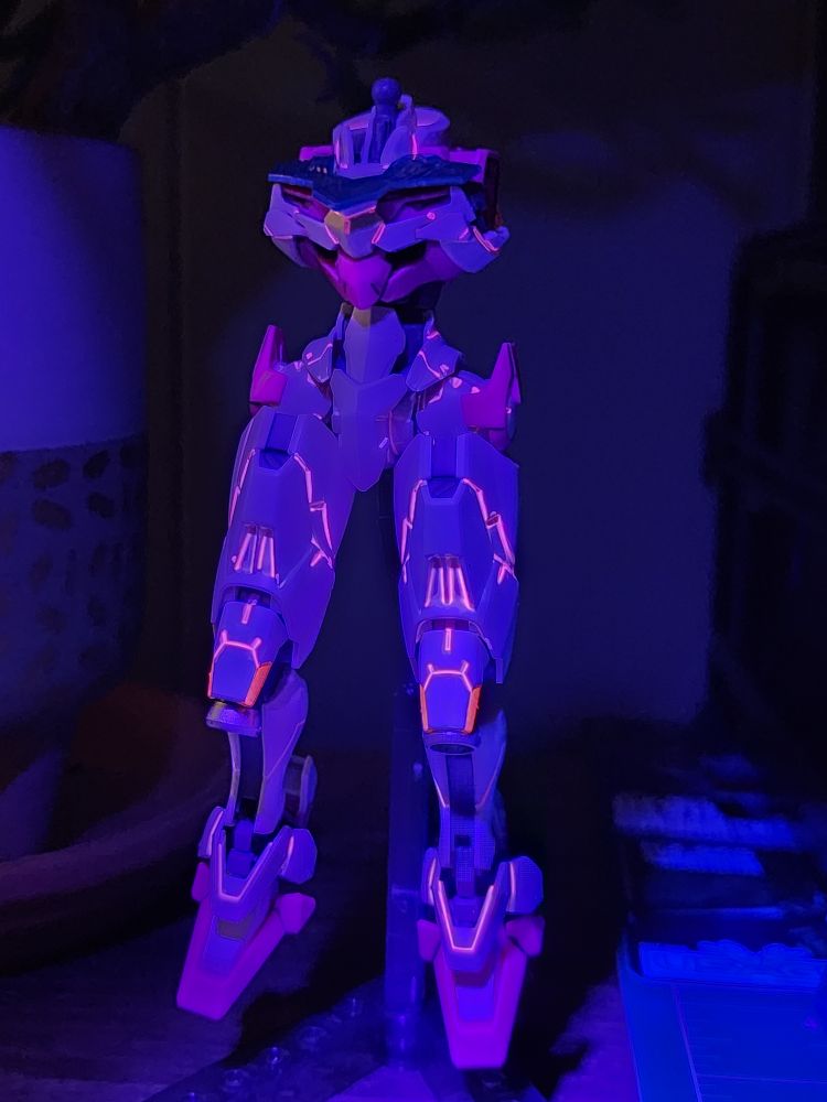 A partially built HG Lfrith model kit from Mobile Suit Gundam: The Witch from Mercury. The legs, waist unit, and chest has been assembled and are on an action base in a relaxed pose facing fhe camera. A black light is shining on the kit to show off fhe fluorescent pink panel lining.