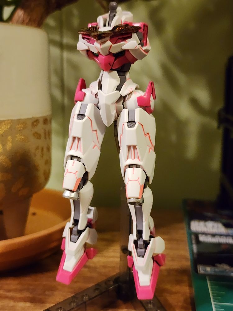 A partially built HG Lfrith model kit from Mobile Suit Gundam: The Witch from Mercury. The legs, waist unit, and chest has been assembled and are on an action base in a relaxed pose facing fhe camera. The kit is panel lined with pink fluorescent panel liner from Stedi.