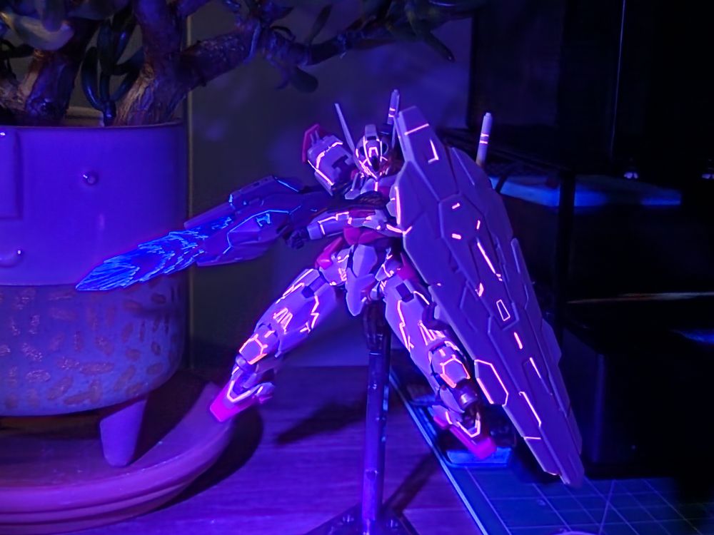 A photo of a completed HG model kit of Gundam Lfrith from Mobile Suit Gundam The Witch from Mercury. The kit is in an action pose blocking with its shield in its right hand and beam rifle in its left. The photo is taken with black showing the flourescent pink and blue Stedi panel liner 