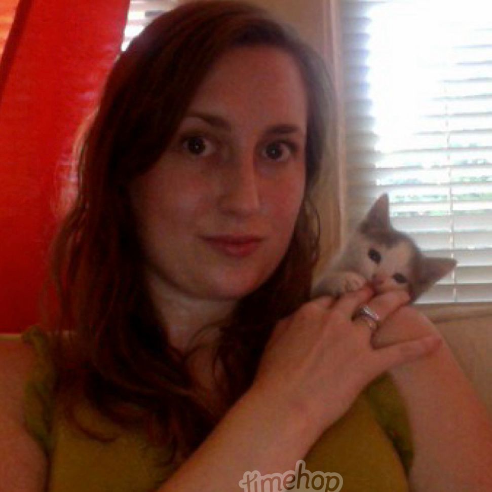 Selfie of a long haired Sarah with a gray and white kitten on her shoulder, attacking her fingers.