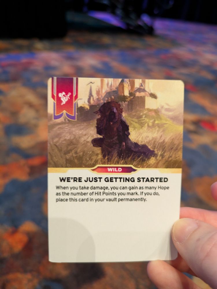 The author holds up a Daggerheart wild card with the title "We're Just Getting Started" against a colorful patterned carpet background.