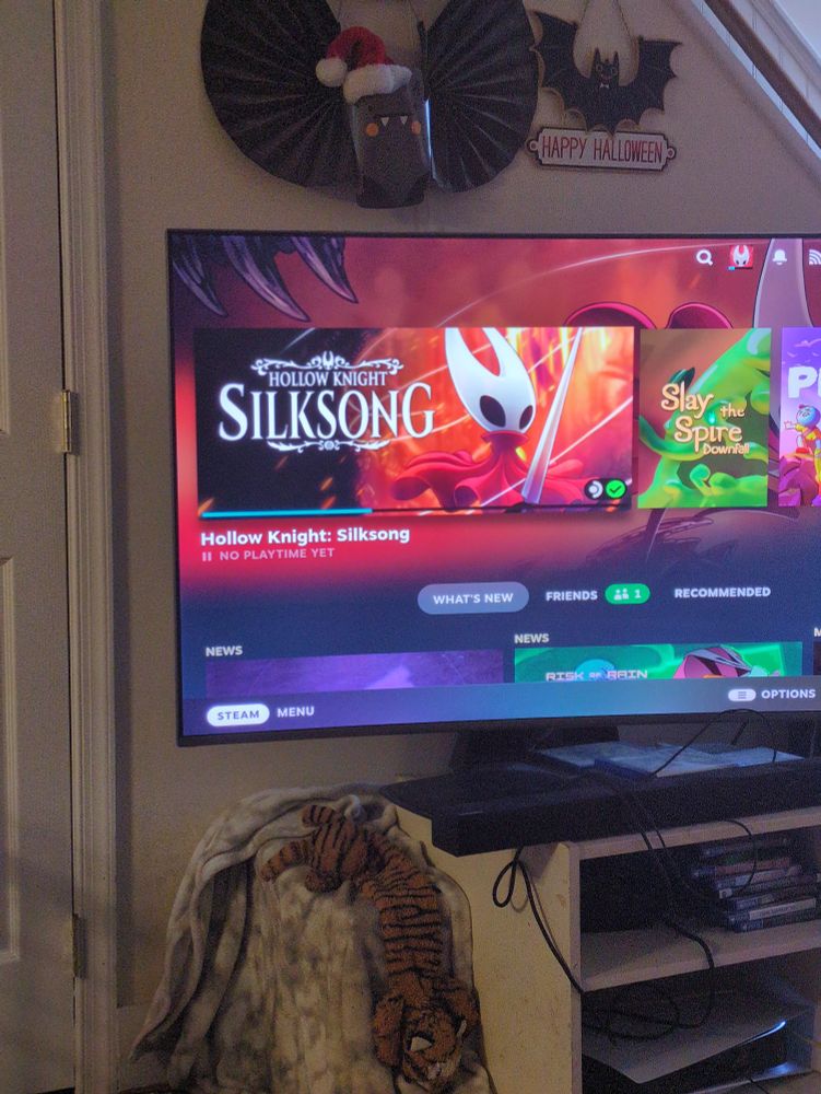 Picture of a curved tv showing the Steam deck interface. The game Silksong is downloading, a progress bar showing at the bottom.