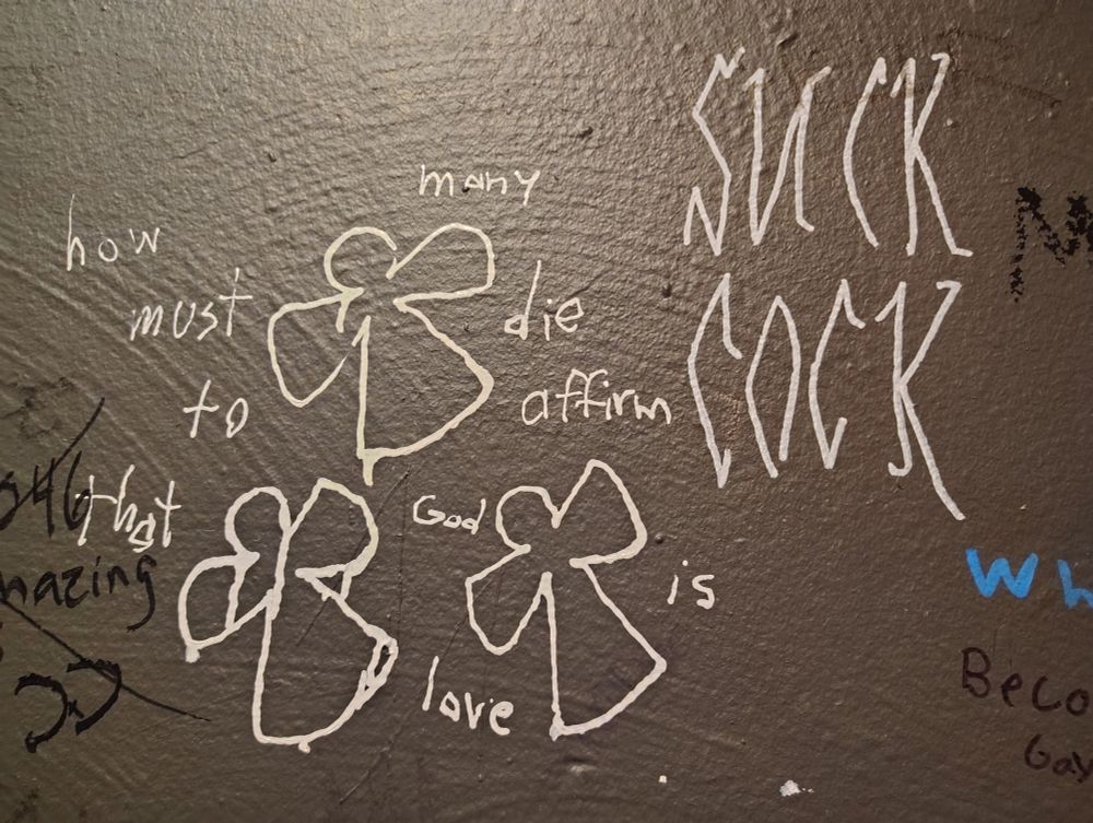 A wall of graffiti. On the left is three crosses with the words "how many more must die to affirm that God is love?" And on the right it says "SUCK COCK"