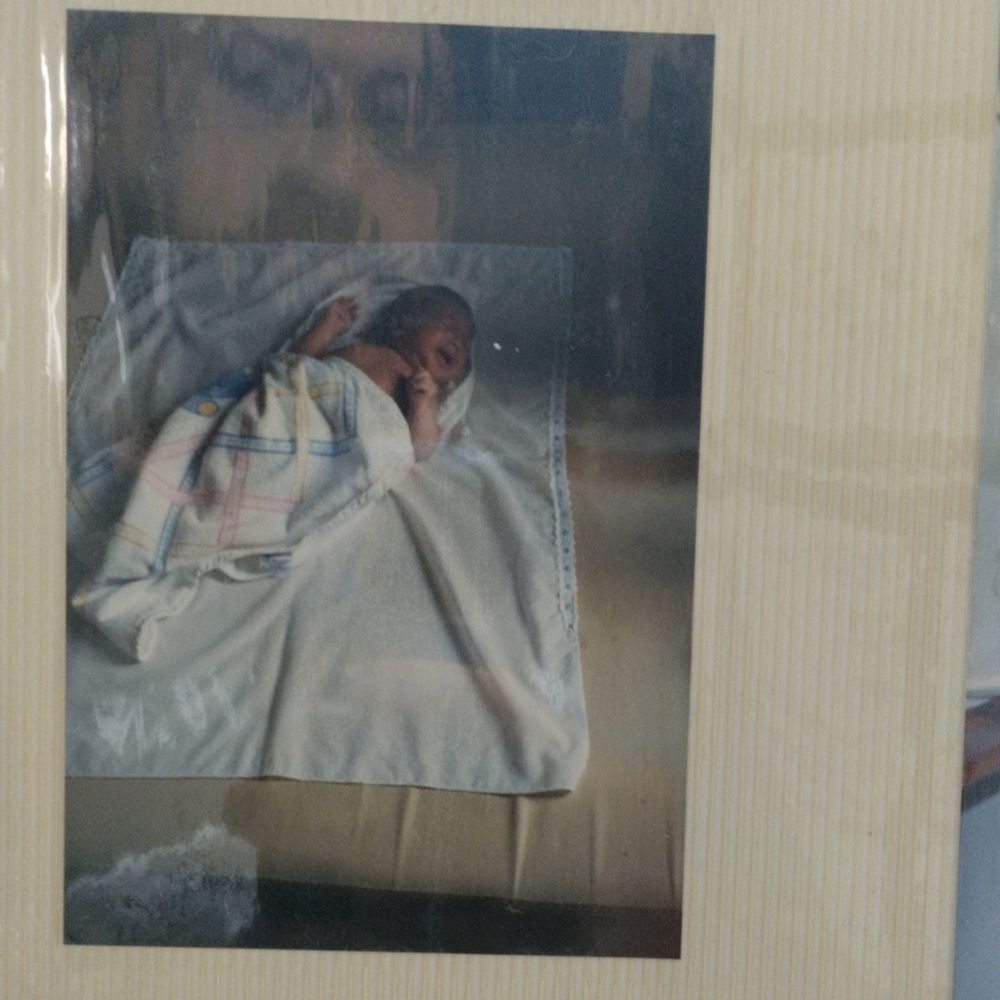 Foto de eu bebê coberto por uma coberta até o peito, com a boca aberta e os olhos fechados. A foto está dentro de um plástico e o meu rosto atual aparece refletido no plástico, ficando acima do bebê, numa parte mais escura da foto