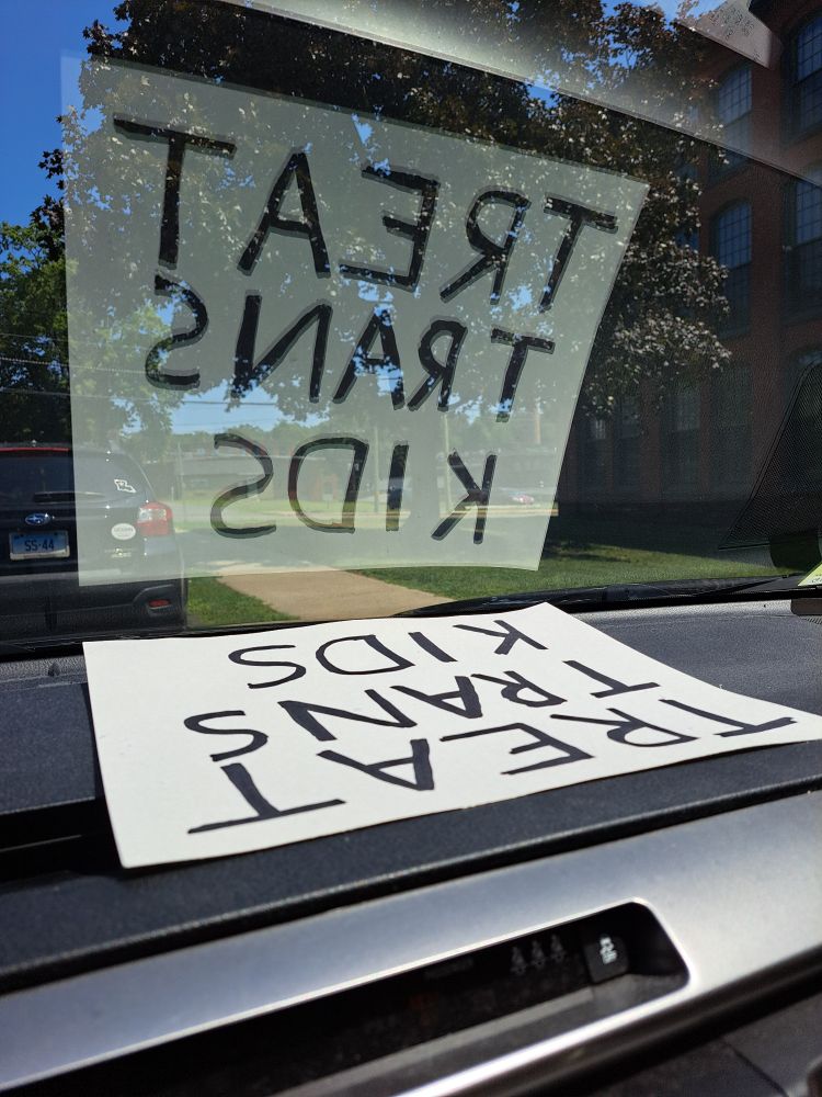 Poster reading "TREAT TRANS KIDS" on the dashboard of a car with reflection in windshield showing 