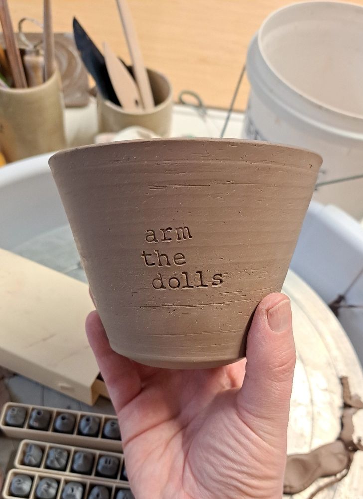 Unfired ceramic mug with "arm the dolls" stamped on it