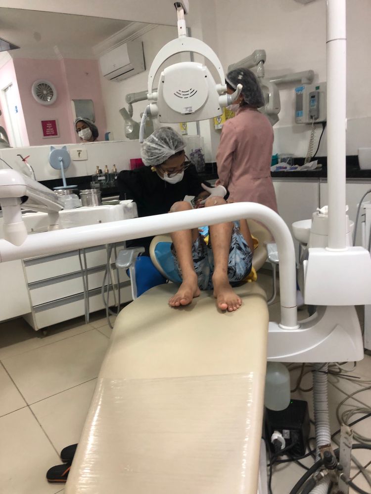 criança deitado na cadeira do dentista com as pernas dobradas. com os braços dobrados tentando segurar a mão da dentista enquanto ela olha dentro da boca dele.