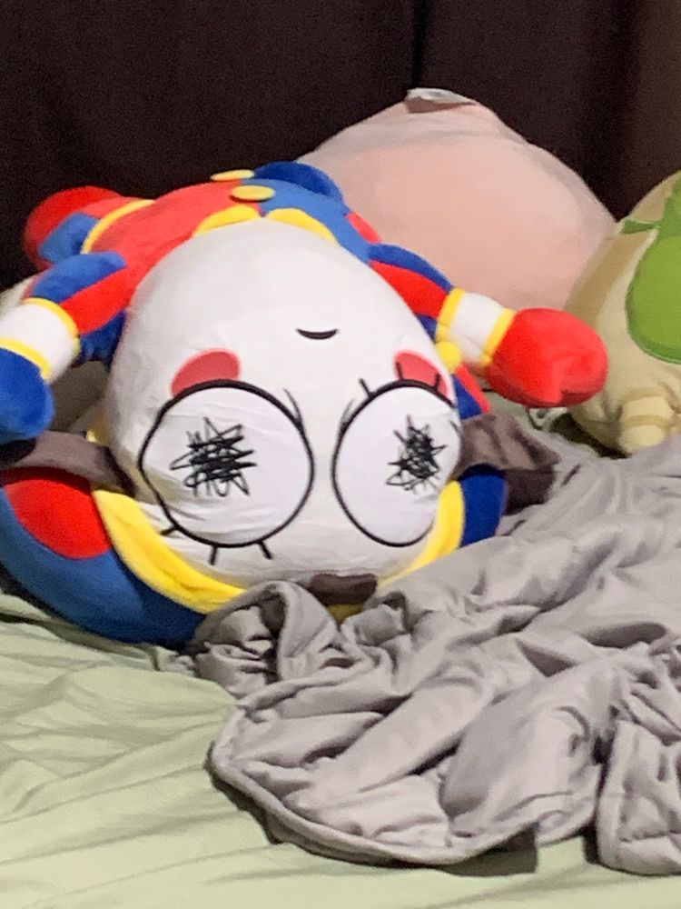 A big pomni plush lying upside down on an unmade bed. Its big ass chin is on full display. 