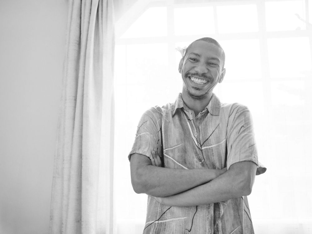 A black an white photo of me smiling. The background is sheers over windows with soft sunlight coming out. Theres a flower in my ear. Im a black person with a buzz cut.
