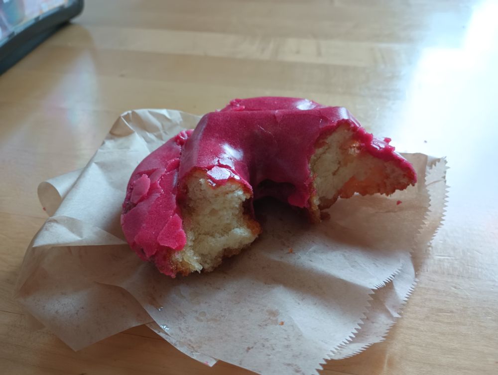 A raspberry-iced old-fashioned donut rests on a greasy napkin. The donut has several large bites taken out of it.