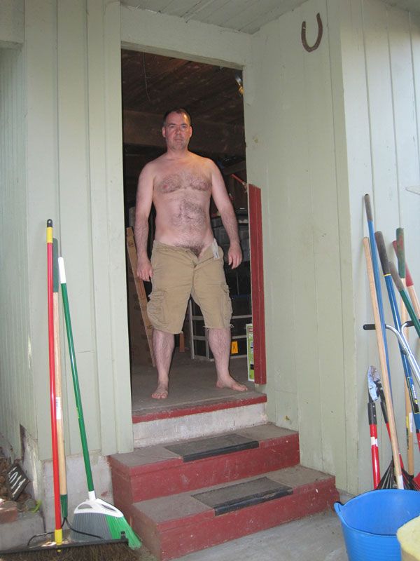 I'm standing in the doorway of the garage at my previous house. There are two steps leading up to the garage and on either side of the door are brooms, yard tools, and some pails. A horseshoe, left by the previous owner, hangs high on the wall.

I'm wearing cutoffs and nothing else. The pants are unbuttoned and the fly is open. I don't have a beard and my hair is short. I'm 50 years old in this photo.