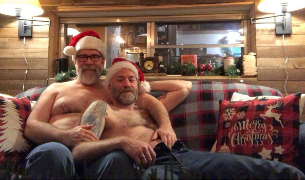Two shirtless, hairy chested guys, wearing Santa hats and relaxing on a couch in a cabin-like setting. There are holiday pillows and decorations around them.