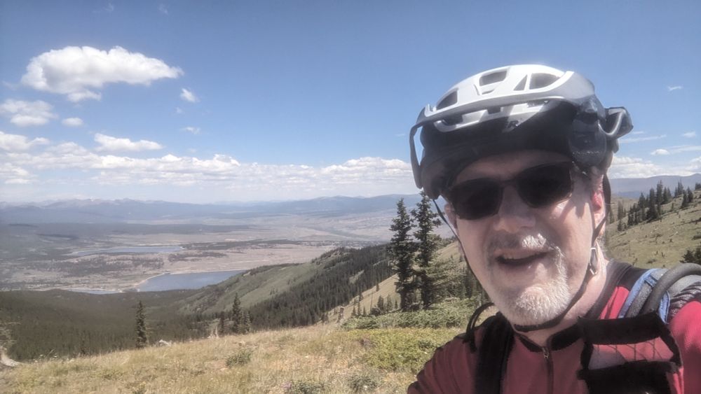Selfie of a cyclist in a helmet and sunglasses with a background looking down over Twin Lakes Colorado, which is far below.