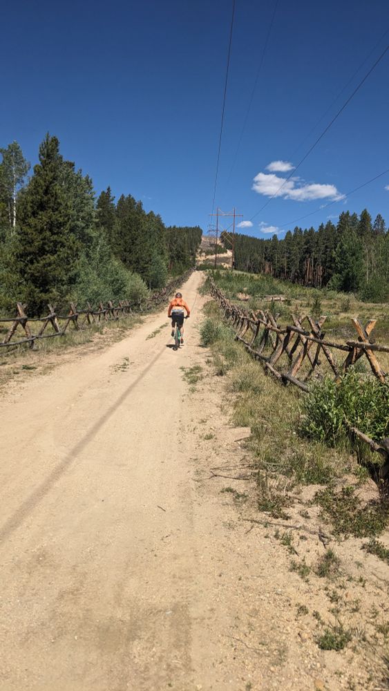 Biker at the bottom of a dirt road heading up a steep hill beneath a power line that goes straight up the slope without any switchbacks.