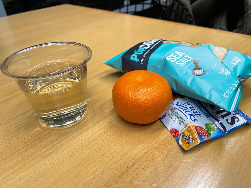 A wooden table with a cup of white wine, a clementine, a bag of pop corners sea salt flavor, and Welches fruit snacks.