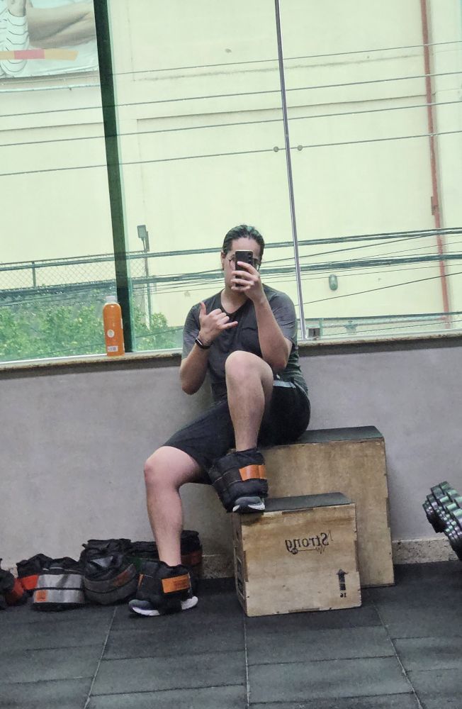 Eu gatinho tirando foto no espelho da academia, com sacos de areia de 5kg nos pés e fazendo o sinal do hangloose 🤙