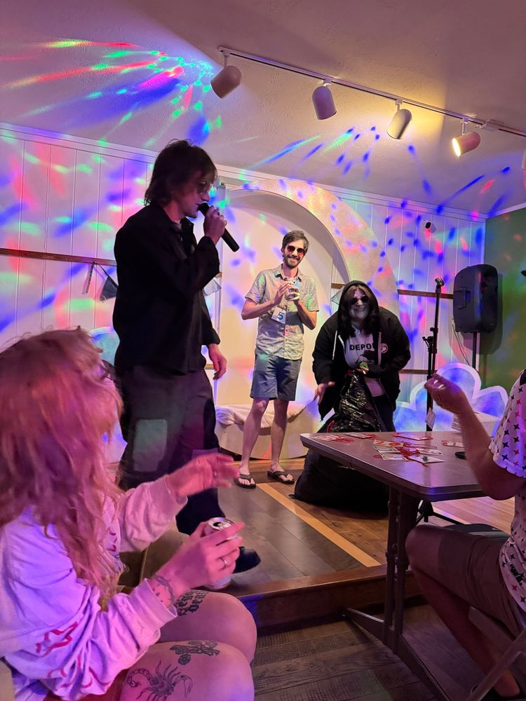 Connor Oberst wears a black shirt and jeans on a small stage amidst colorful lights. The winning  competitor is laughing behind a heaping bag of his old clothes. 