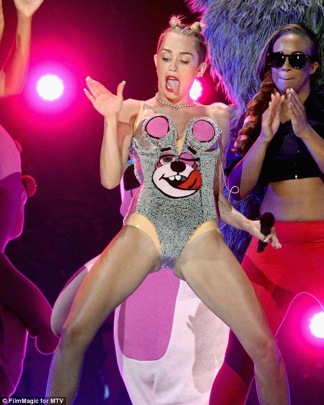 Miley Cyrus in mouse licking face and winking leotard. She is licking downwards. The mouse's ears cover her breasts. 