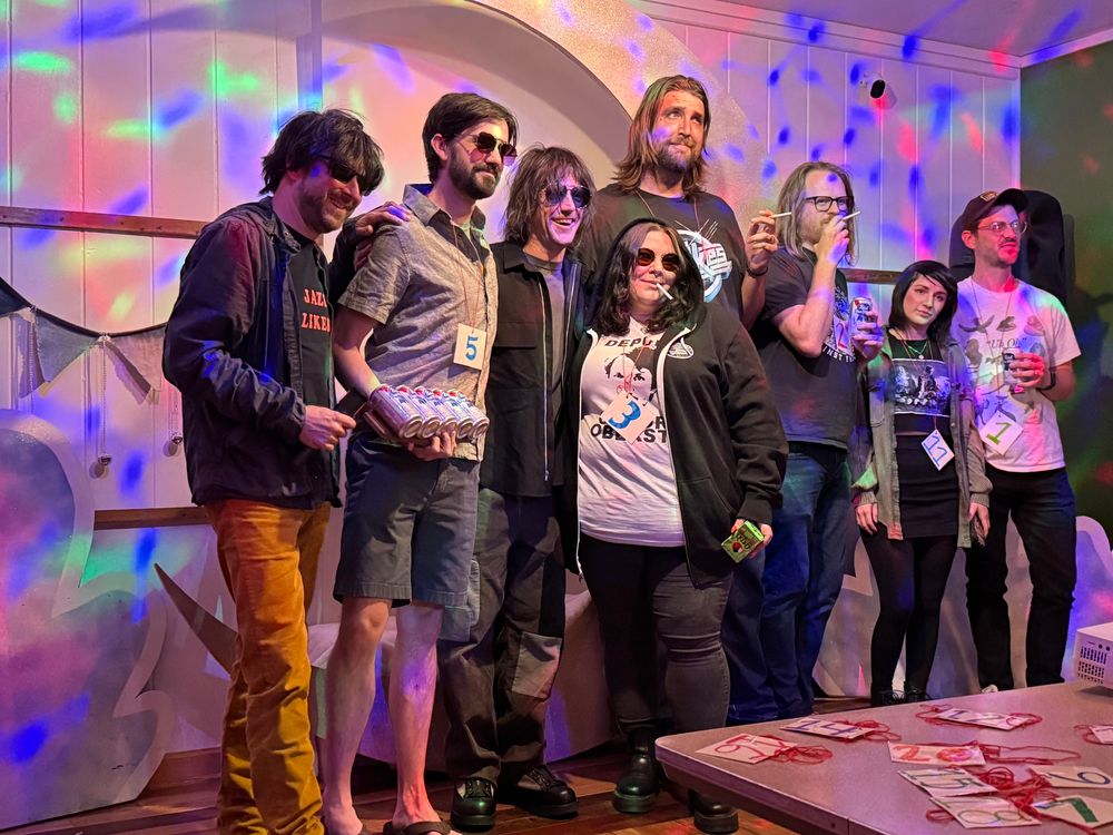 Group picture of eight lookalike competitors posing with Connor. Colorful lights, wooden stage. Lots of cigarettes and shades. 