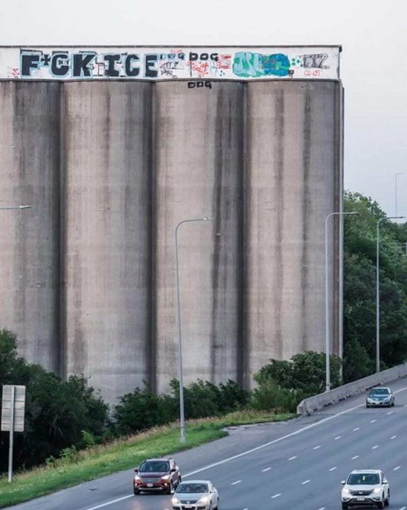 Concrete silos just south of the busiest stretch of Interstate 80 in Omaha. The top structure is frequently tagged with graffiti. It currently features big black letters with light blue outlines that read F*CK ICE