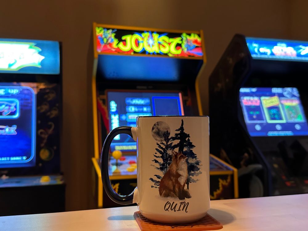 3 arcade machines behind a mug with artwork on it of a fox sitting in front of a pine tree under the moon, the mug is on a Vans coaster on a bar table. 