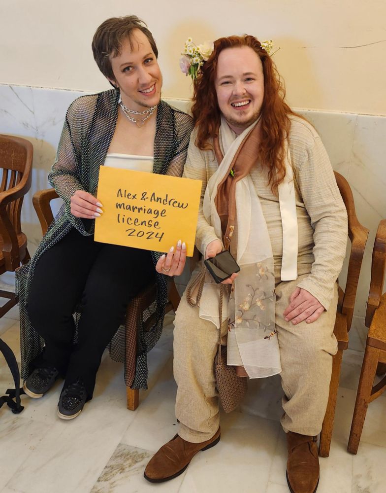 A photograph of Alex and Andrew smiling and holding their marriage license in a manila envelope that says "Alex & Andrew marriage license 2024" Alex is a light skinned person of Russian, Syrian, and European descent with short brown hair. He is wearing a sparkly duster jacket, a cream tube top, black pants, sparkly shoes, and a heart chain necklace. Andrew is a light skinned person of white European descent with long wavy red hair with an arrangement of pink and white flowers woven into his hair. He is wearing a cream colored silk shirt and pants, and a long silk scarf with cream and rust colors and embroidered flowers. 