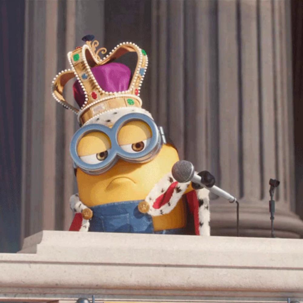 a minion wearing a crown and a king 's robe stands at a podium with a microphone