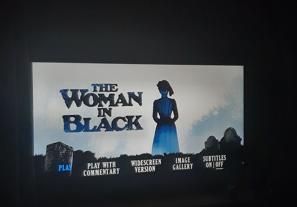 A tv showing the menu screen for 1989 ITV version of THE WOMAN IN BLACK. The titular woman, in a black dress and hat, is stood in a graveyard, staring at the camera.