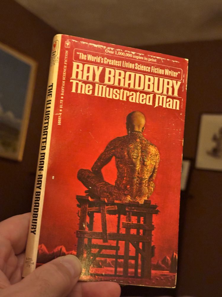 Photo of a paperback copy of Ray Bradbury’s, “The Illustrated Man.” The cover depicts a naked man with shaved head sitting cross legged, facing away from the viewer, on a short and rickety wooden platform. His body is completely covered in tattoos, right up to his neck.