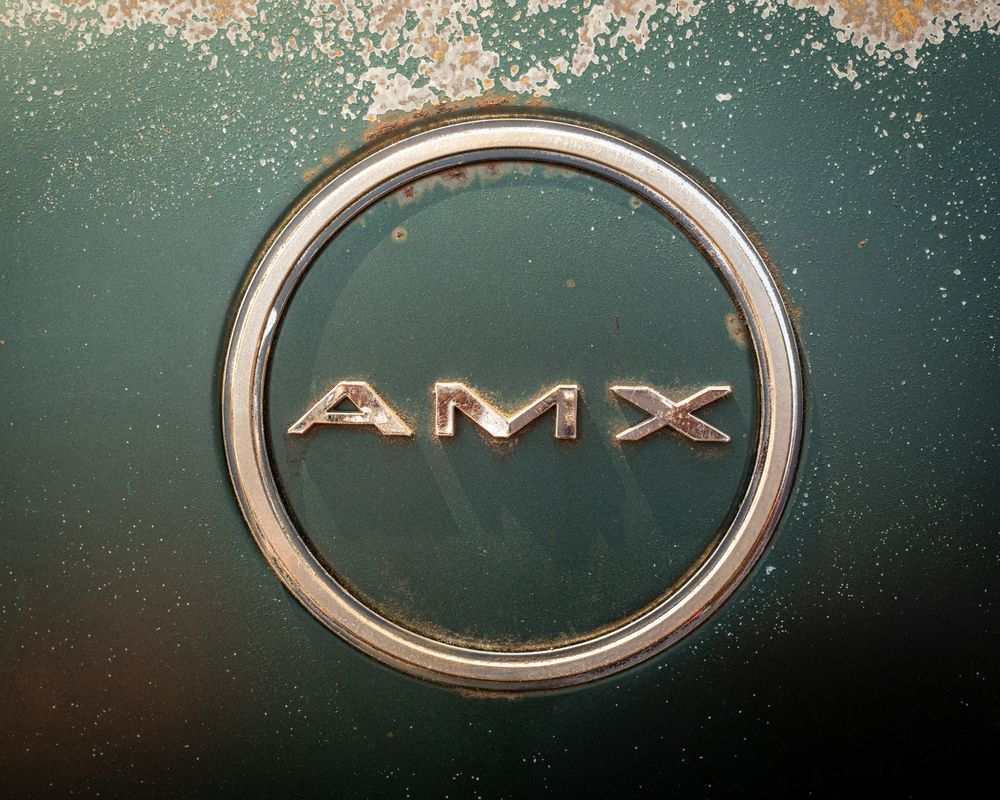 Color photo of the AMX badge on a green AMC Javelin AMX. The badge and surrounding body paint are pitted and show signs of rust.