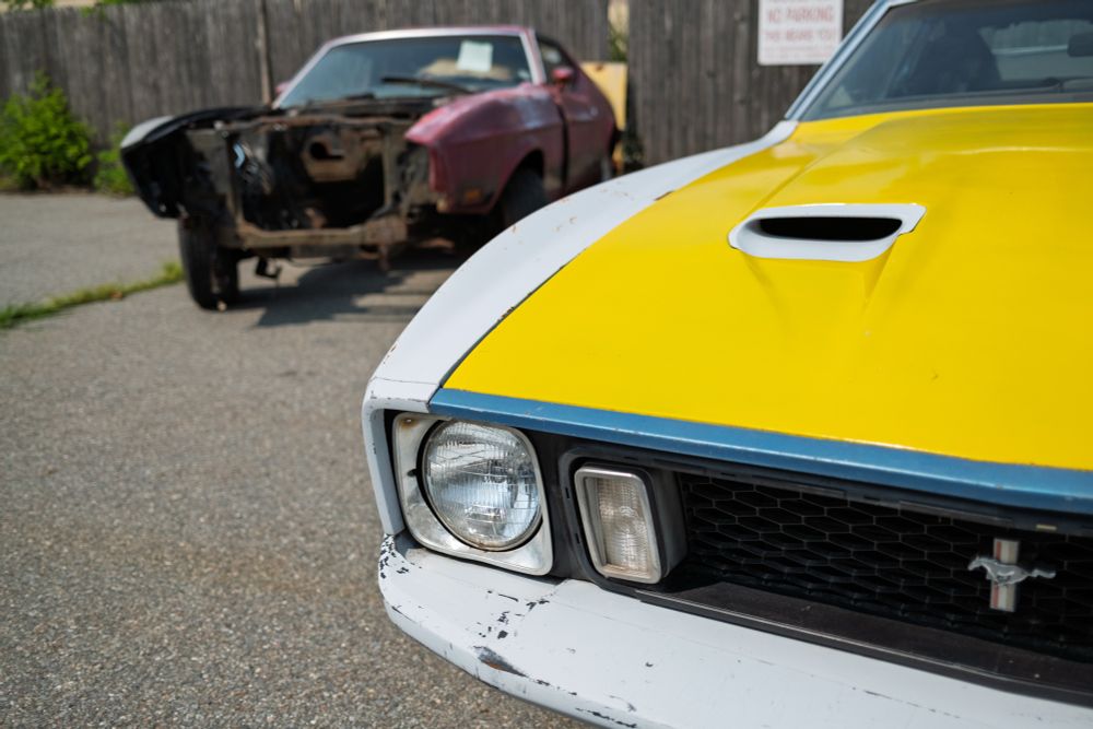 Color photo of the front left side of a early 70’s Mach1 Ford Mustang. It has a bright yellow hood with a thin blue front edge, a white bumper and fender. Just beyond is the hulking mess of another Mach1 that lacks its grille, bumper, radiator, and engine.