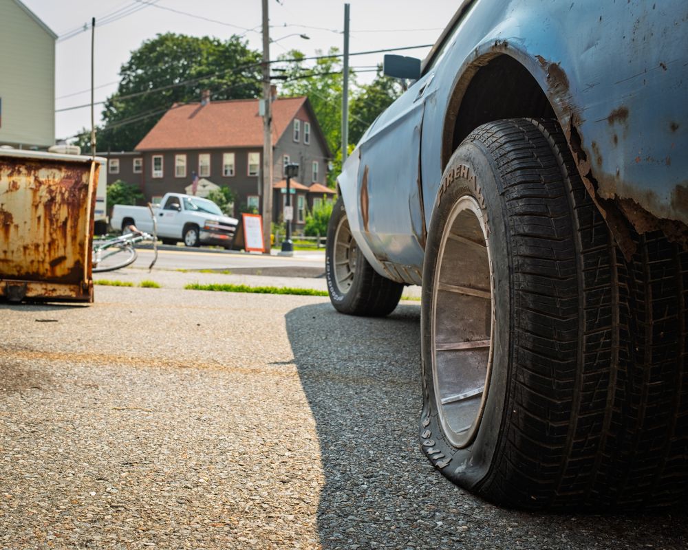 Color photo of a used car/mechanic’s lot featuring the left side of a Mach 1 Mustang from the rear… it has a flat tire. There’s some kind of rusted metal container to the left, a bicycle and pickup truck in the middle distant background.