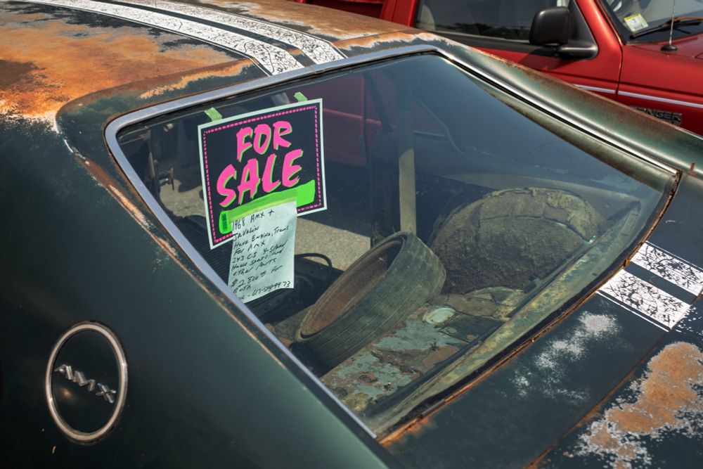 Read window of an AMC Javelin AMX showing a rusted rooftop with dual white racing stripes down the middle. Body color is dark green. The circular AMX badge is visible in the lower left and a FOR SALE sign is hung inside the window.