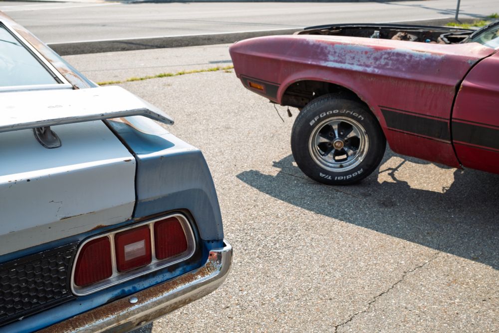 Color photo of the right rear of a Ford Mach1 Mustang showing its tail light and rear spoiler. The spoiler and trunk lid are white, the rest of the visible part of the car’s body is light blue. The chrome bumpers shows quite a bit of rust. To the right of that car is the driver’s side front of another Mach1 Mustang showing its red driver’s side fender and door. The car looks lifted up pretty high because it has no engine to weigh down its shock absorbers.