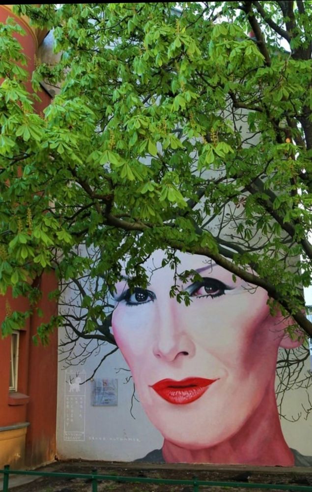 Großformatiges Porträt einer Frau mit ausdrucksstarken Augen und Lippen in kräftigem Rot. Ein Baum mit dichten, grünen Blättern steht direkt vor der Wand. Die Zweige und Blätter wirken wie das Haar der Frau im Bild. 