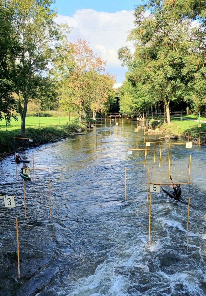 Abschnitt eines Wildwasser-Kanuslalom-Kurses in einer natürlichen Flussrinne. Entlang des Flusses hängen zahlreiche Tore – das sind nummerierte Schildchen an Stangen mit quer darüber gespannten Stäben. Die Tore sind teilweise grün/weiß oder rot/weiß. Im Wasser befinden sich drei Kanusportler in schlanken Slalomkajaks. Das Wasser ist klar, hat leichte bis mittlere Strömung und bildet im Vordergrund weiße Wellen.
Beidseits des Flusses stehen dichte, grüne Bäume. Der Himmel ist blau mit einigen weißen Wolken.